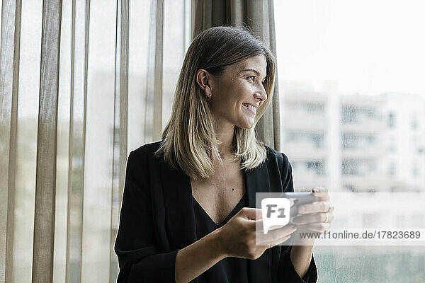 Smiling businesswoman holding mobile phone looking through window