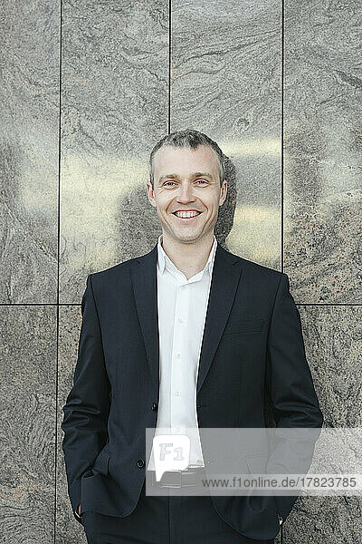 Happy businessman with hands in pockets in front of wall
