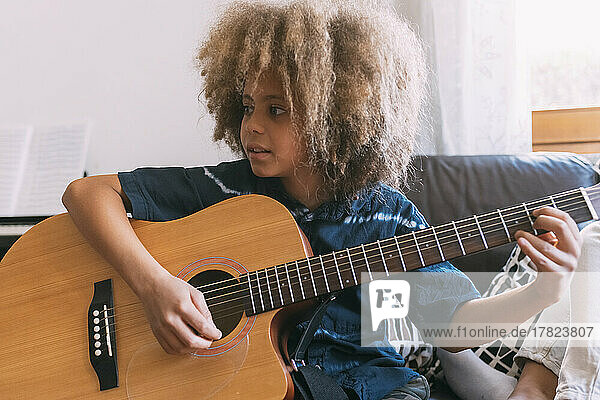 Boy with Afro hairstyle playing guitar sitting on sofa at home
