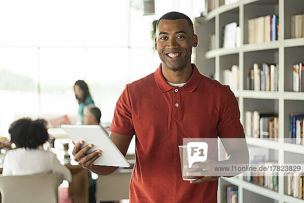 Smiling businessman holding disposable cup and tablet PC standing at office