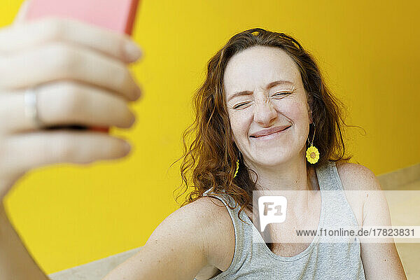 Happy woman with eyes closed taking selfie through mobile phone in front of yellow wall