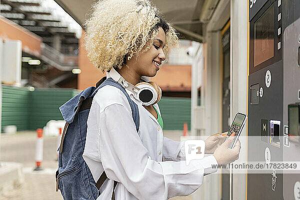 Smiling woman scanning QR code through smart phone on ticket vending machine