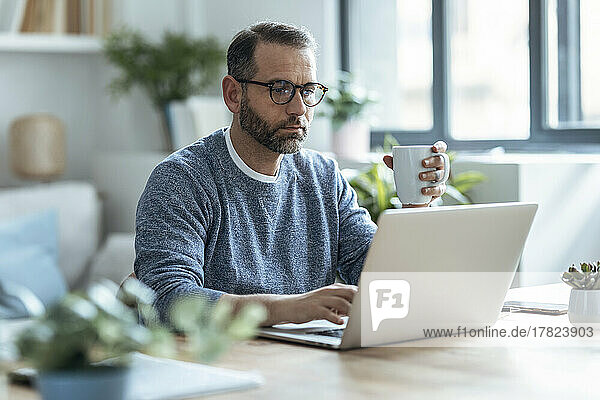 Businessman holding coffee mug working on laptop at home