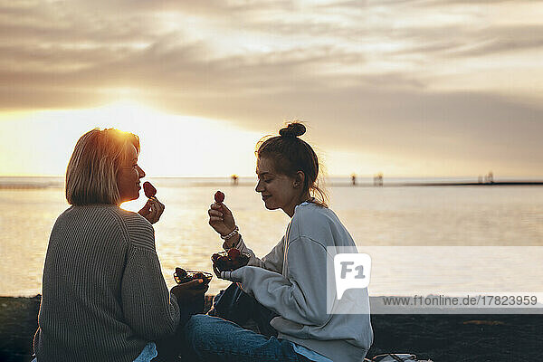 Smiling mother and daughter eating strawberries at beach on sunset