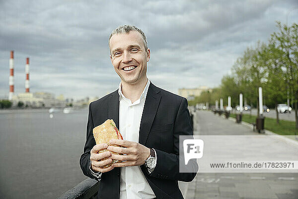 Smiling businessman with sandwich standing on promenade