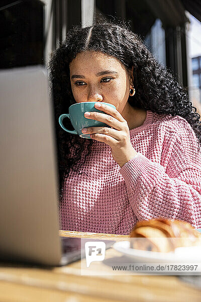 Young woman drinking coffee using laptop at sidewalk cafe