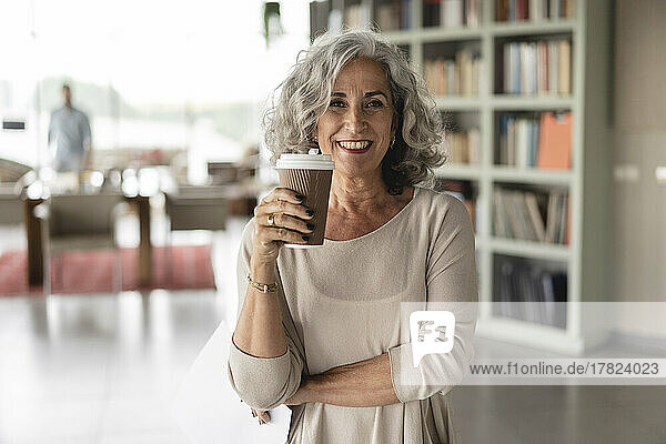 Senior smiling businesswoman holding disposable cup standing at office