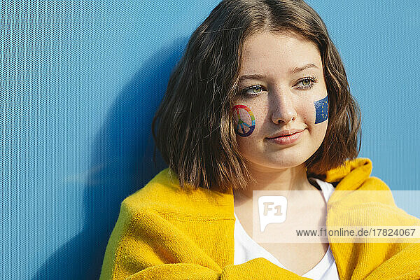 Thoughtful teenage girl with peace symbol and European Union paint on cheeks in front of blue wall