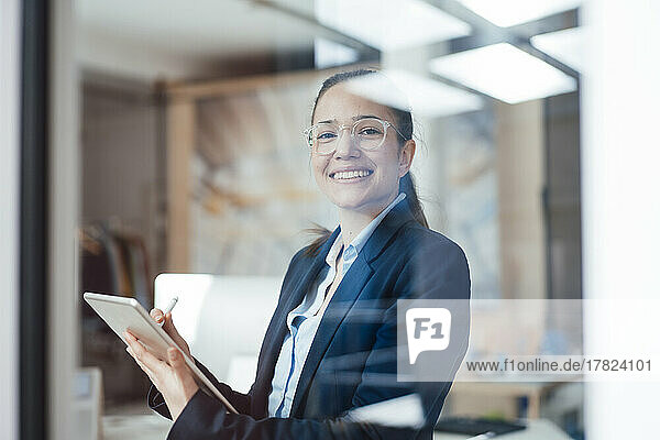 Happy businesswoman with tablet PC seen through glass of office