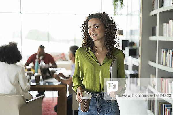 Contemplative businesswoman with laptop and disposable cup standing at office