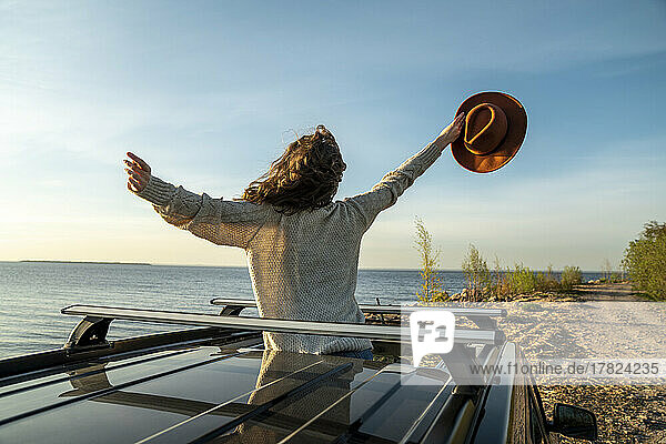 Young woman holding hat standing with arms outstretched through sun roof of car