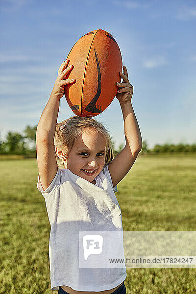 Glückliches blondes Mädchen trägt Rugbyball auf dem Kopf auf dem Sportplatz