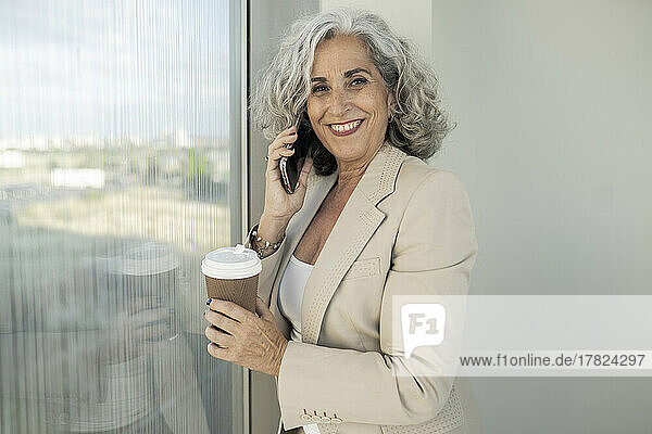 Smiling businesswoman talking on smart phone holding disposable cup