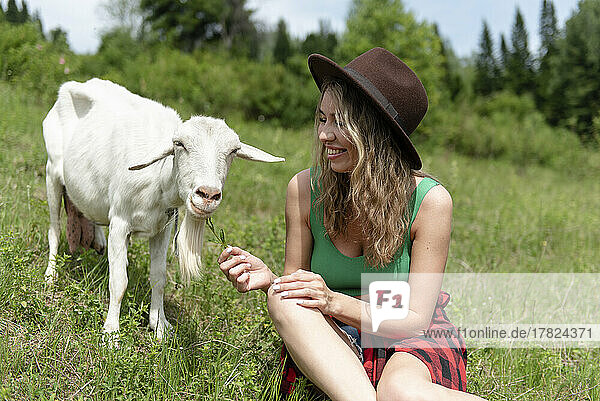 Smiling woman sitting on grass and feeding goat