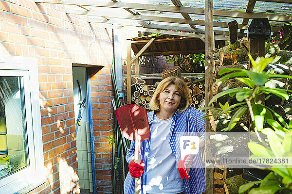 Lächelnde Frau mit Gartenschaufel steht am Schuppen