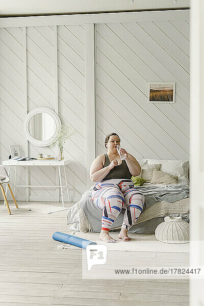 Frau trinkt Wasser aus einer Flasche und sitzt zu Hause im Bett