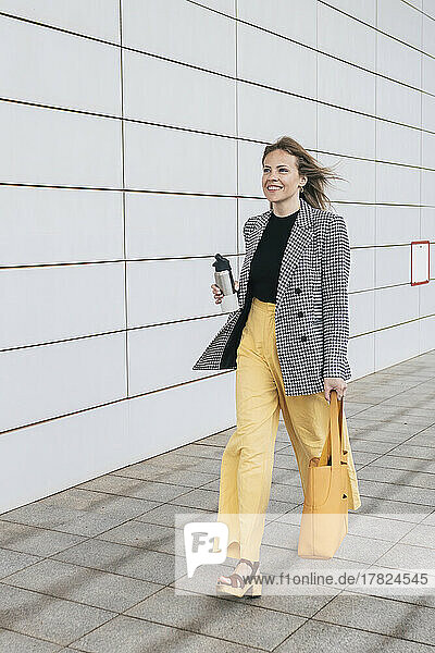 Businesswoman carrying bottle and bag walking by wall