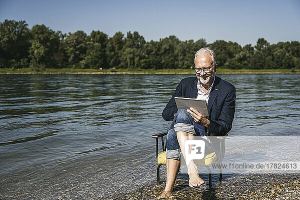 Lächelnder älterer Mann sitzt mit Tablet-PC auf einem Stuhl am Flussufer
