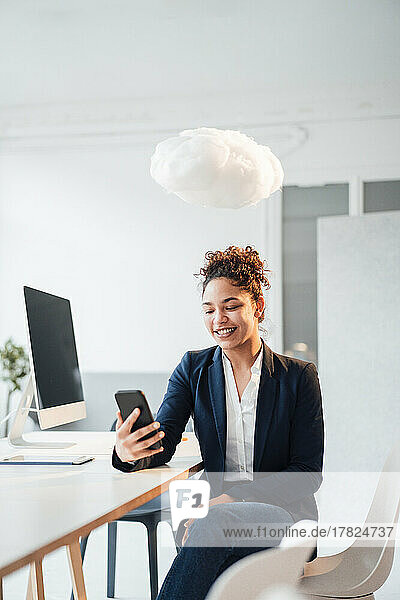 Wolke schwebt über glücklicher Geschäftsfrau  die im Büro ihr Smartphone benutzt