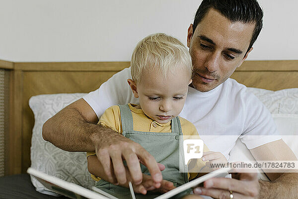 Man reading book with son sitting on bed at home