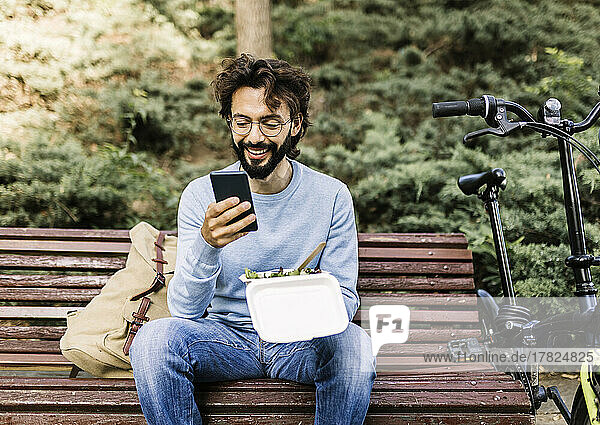 Smiling man with lunch box using phone sitting on bench