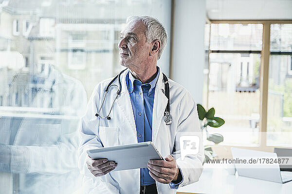 Thoughtful doctor with tablet PC looking through window in office