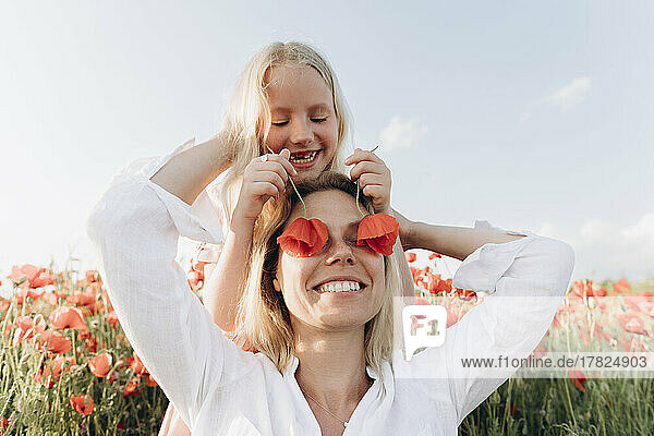 Happy girl hiding mother's eyes with flower in poppy field