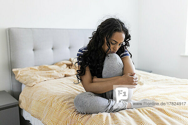 Young woman hugging knee on bed in bedroom