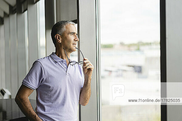 Thoughtful businessman holding eyeglasses looking out of window at office