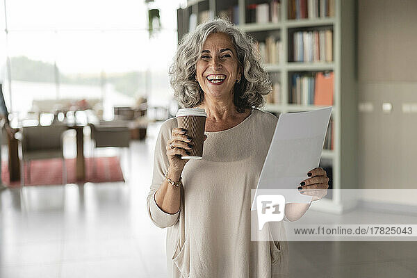 Happy businesswoman holding disposable cup and document standing at office