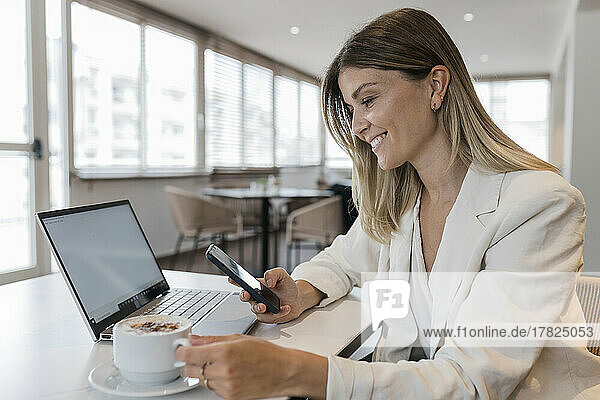 Smiling businesswoman holding mobile phone and coffee cup sitting at cafe
