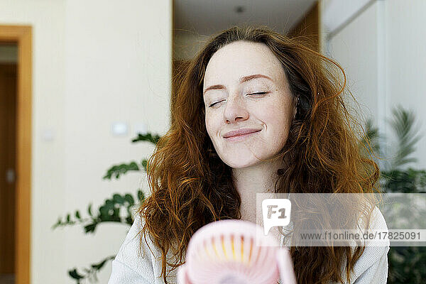 Smiling woman with eyes closed holding portable fan at home