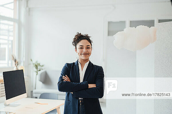 Lächelnde Geschäftsfrau mit verschränkten Armen steht neben einer schwebenden Wolke im Büro