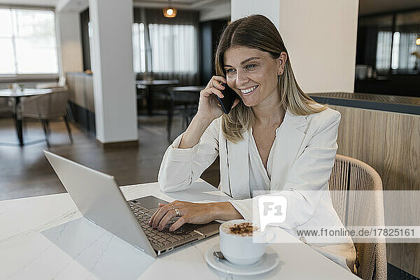 Smiling businesswoman talking on phone sitting with laptop at cafe
