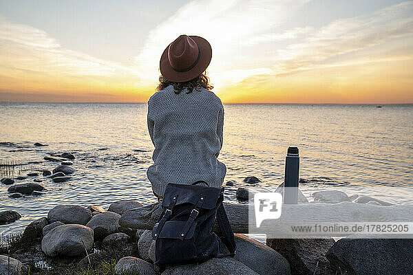 Junge Frau mit Rucksack sitzt bei Sonnenuntergang auf einem Baumstamm
