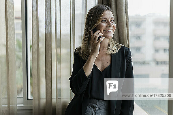 Smiling businesswoman talking on phone standing by window