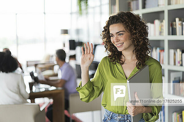 Smiling businesswoman waving holding laptop at office
