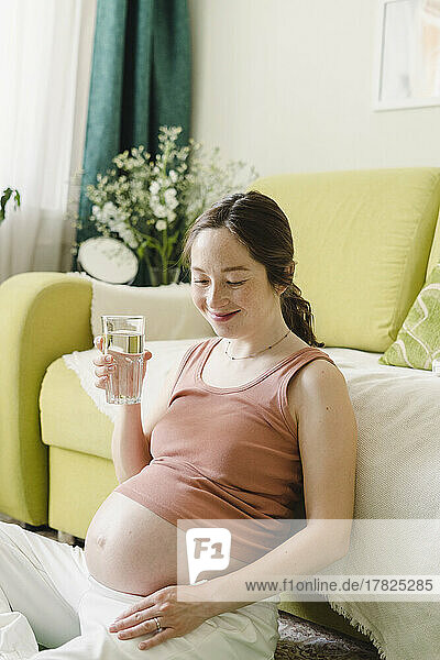 Smiling woman with glass of water sitting by sofa at home