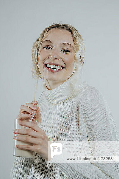 Smiling young woman in turtleneck holding glass of plant milk against white background