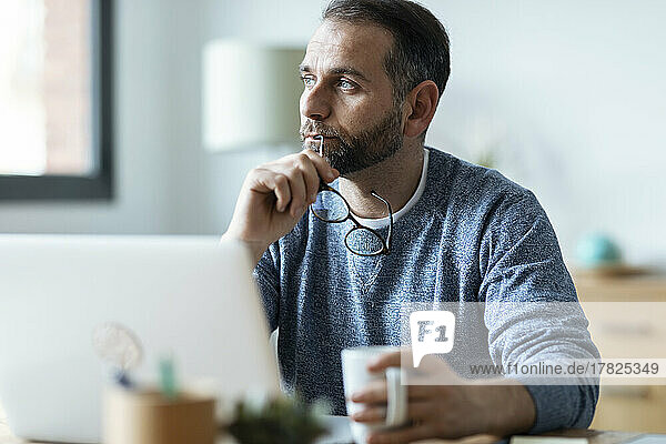 Thoughtful freelancer holding eyeglasses sitting with mug at home