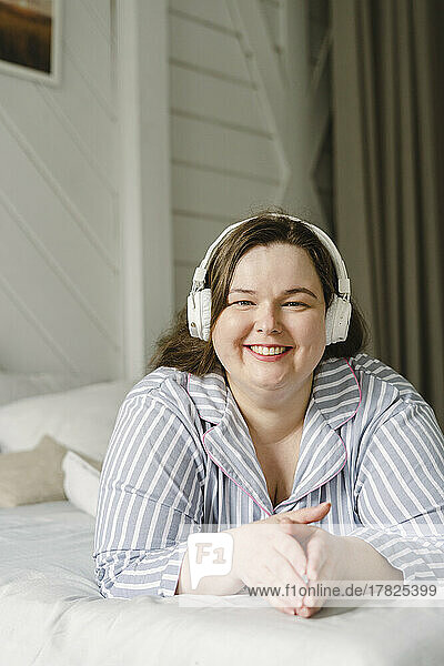 Smiling woman listening music through wireless headphones lying on bed