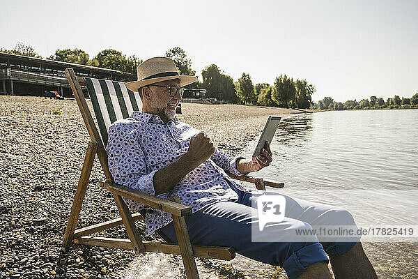 Happy man holding tablet PC sitting on deck chair at riverbank