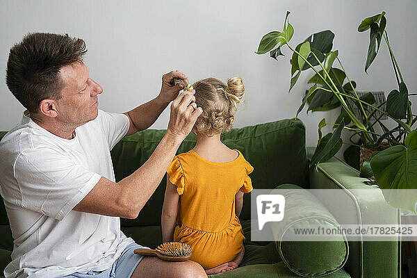 Vater bindet seiner Tochter  die zu Hause auf dem Sofa sitzt  die Haare