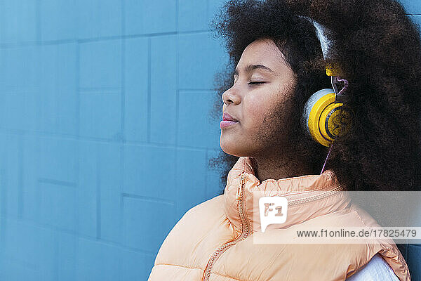 Girl with eyes closed listening music leaning on blue wall