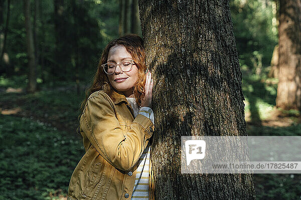Lächelnde Frau mit geschlossenen Augen umarmt Baum im Wald