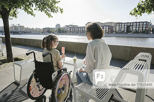 Mädchen mit Behinderung isst Eis und sitzt im Rollstuhl neben ihrer Mutter am Flussufer