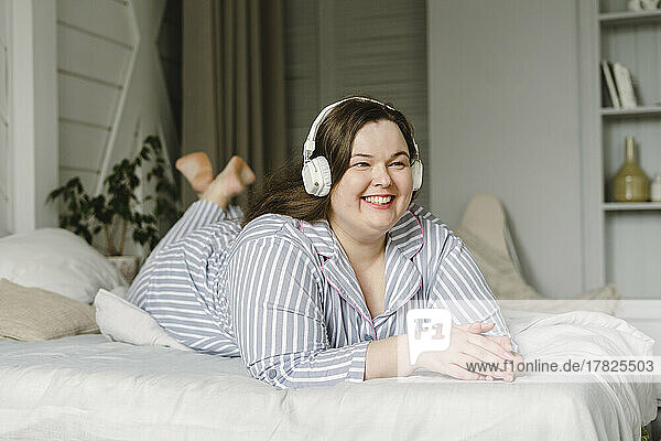 Happy woman enjoying music through wireless headphones lying on bed at home