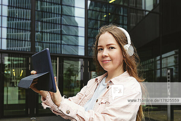 Happy woman wearing wireless headphones standing with tablet PC in front of building