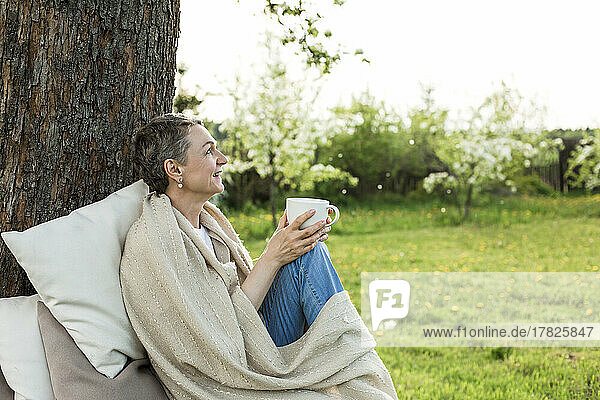 Happy woman wrapped in blanket holding cup sitting on bench around tree trunk