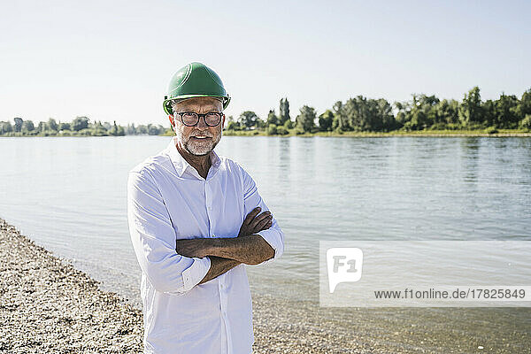 Smiling senior engineer wearing green hardhat standing with arms crossed at riverbank